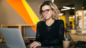 Jovem especialista sorrindo enquanto digita no notebook em um escritório moderno e colorido, estudando o passo a passo de como criar um chatbot para automatizar o atendimento ao cliente.